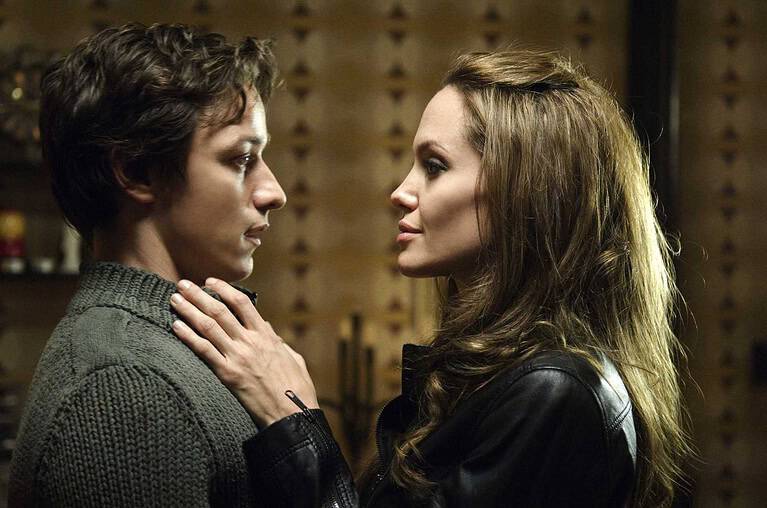 A young man and woman facing each other closely, displaying emotional intensity, in a dimly lit room. the woman gently touches the man's chest.