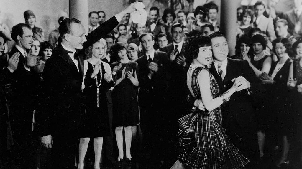 A black-and-white photo of a man and a woman dancing joyfully in front of an applauding crowd at a formal event.