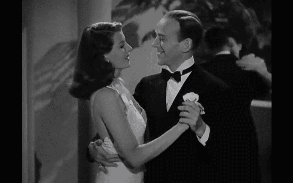 A man in a tuxedo and a woman in a white gown are dancing and smiling at each other in a black-and-white movie scene.