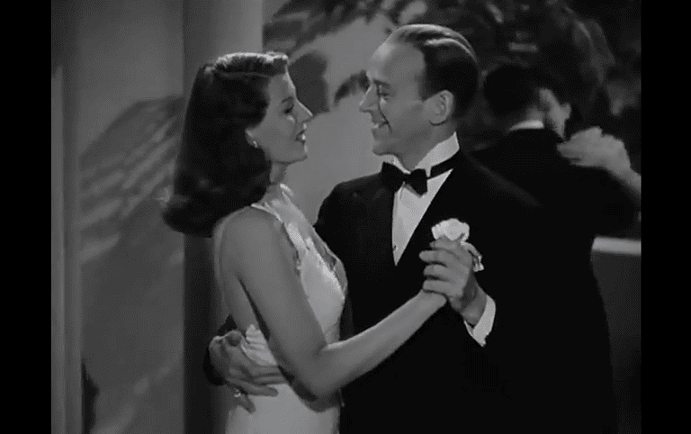 A man in a tuxedo and a woman in a white gown are dancing and smiling at each other in a black-and-white movie scene.
