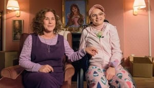 ‘The Act’: The Complete Limited Series Blu-Ray Review – A Disturbing Portrait Of Psychological Treachery Two women sitting and smiling in a cozy room with cardboard boxes and a portrait on the wall. one woman is older and uses a wheelchair, the younger wears a headscarf and glasses.