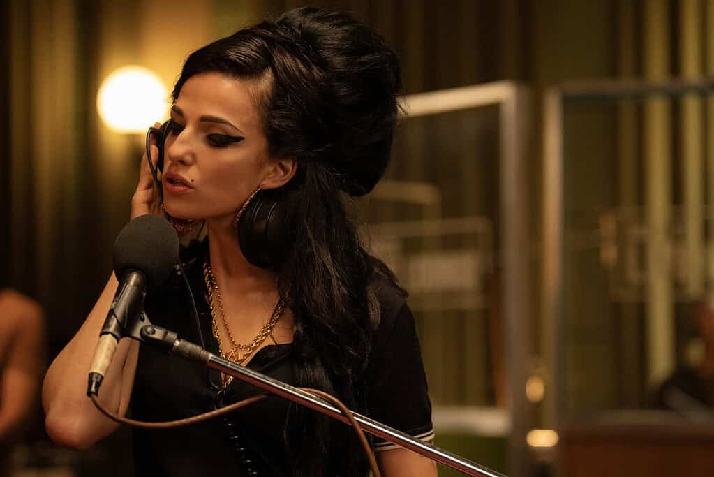 A woman with a dramatic beehive hairstyle sings into a microphone, wearing large hoop earrings and stylish makeup in a warmly lit room.