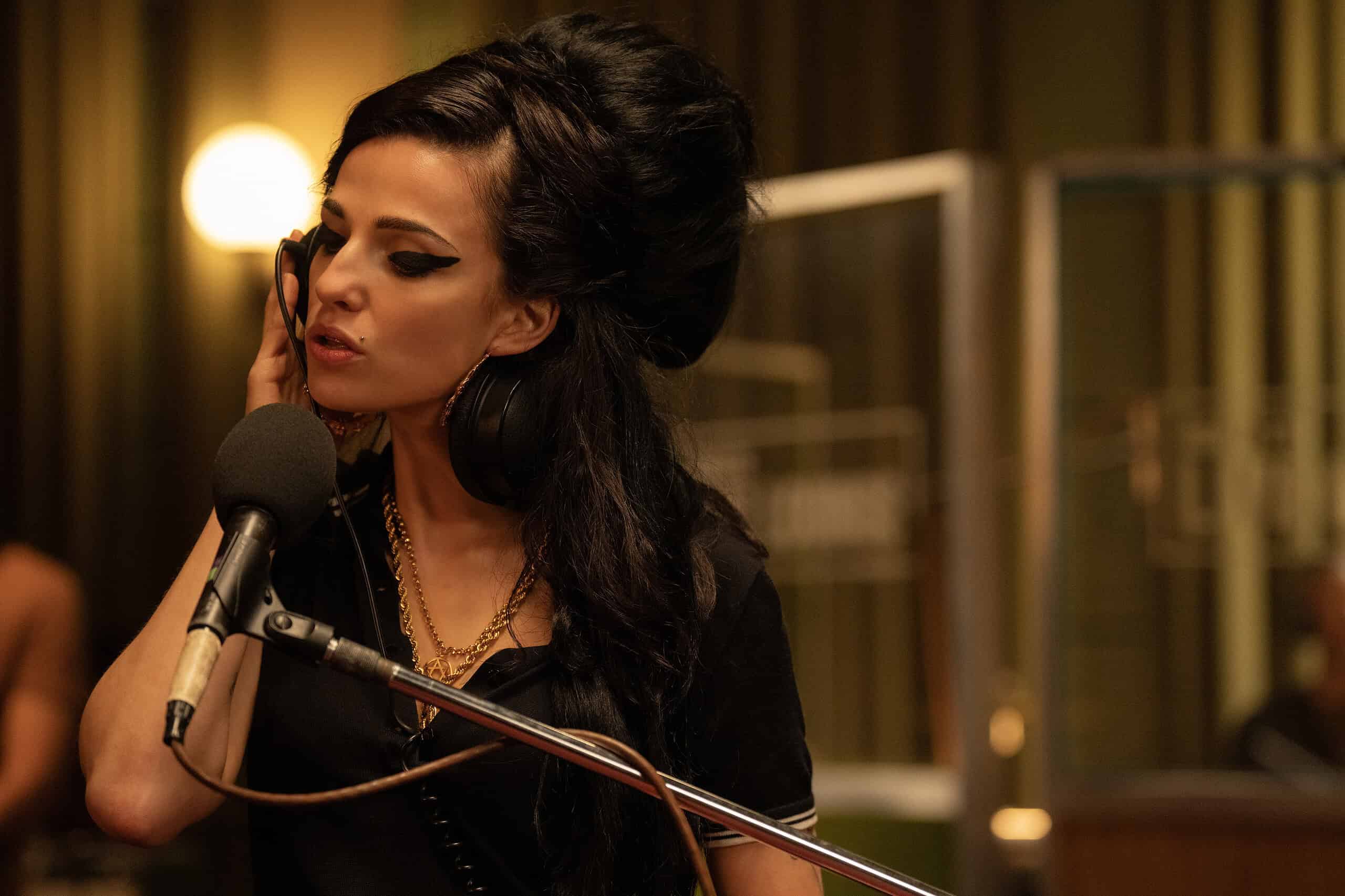 A woman with a dramatic beehive hairstyle sings into a microphone, wearing large hoop earrings and stylish makeup in a warmly lit room.
