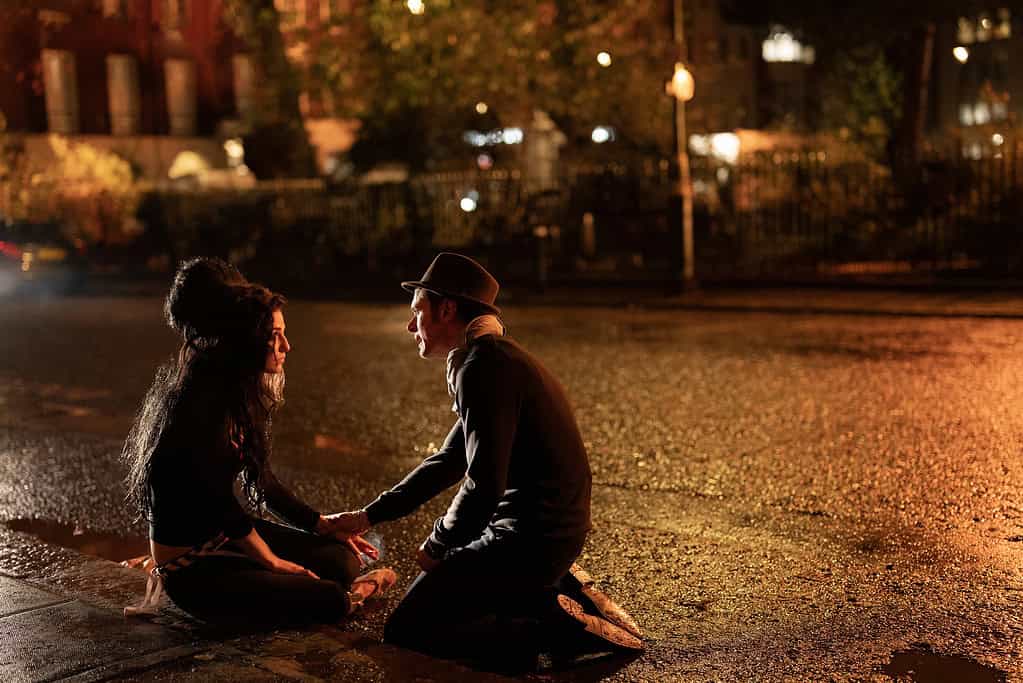 A couple sits on a wet street at night, holding hands and facing each other with a softly lit park fence in the background.