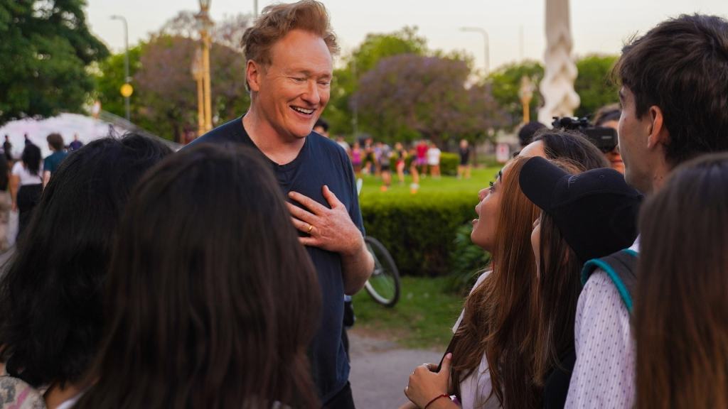 A man with red hair laughing and interacting with a group of young people in a park.