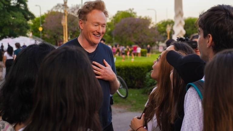 A man with red hair laughing and interacting with a group of young people in a park.