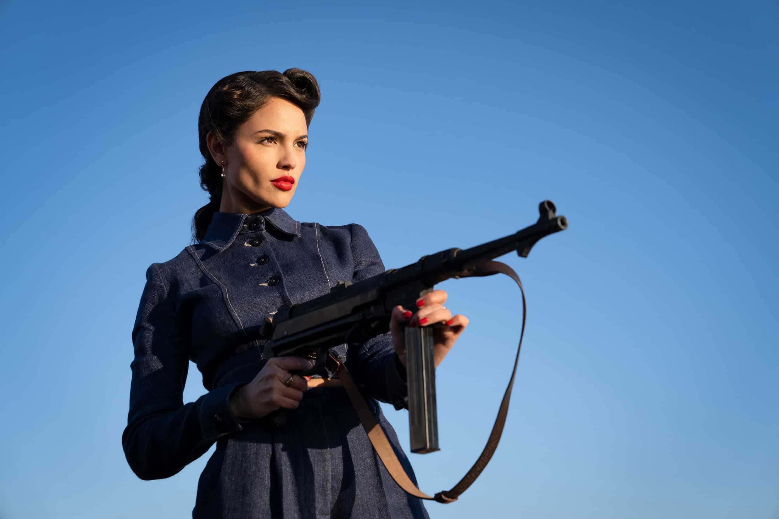 A woman in a vintage military uniform holding a rifle against a clear blue sky.