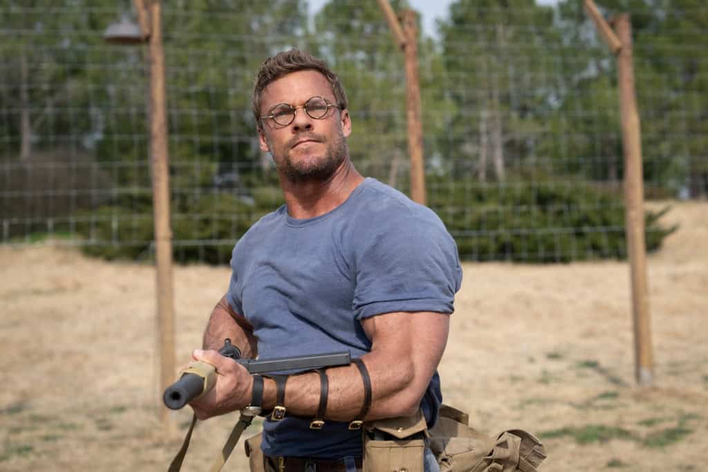 Man wearing glasses and tool belt poses with a sledgehammer in a construction setting, looking determined.