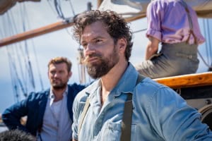 Two men on a sailboat, the foreground showing a bearded man in a blue shirt and suspenders gazing intently, with another man partially visible behind him.