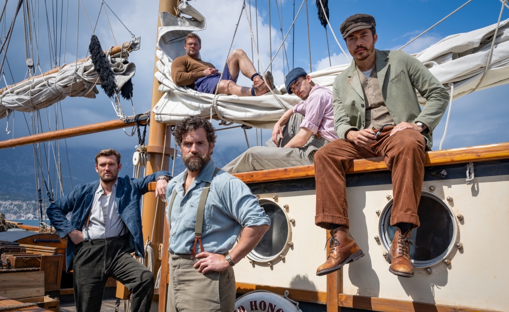 Five men in period clothing pose on the deck of a vintage sailboat, with rugged mountain scenery in the background.