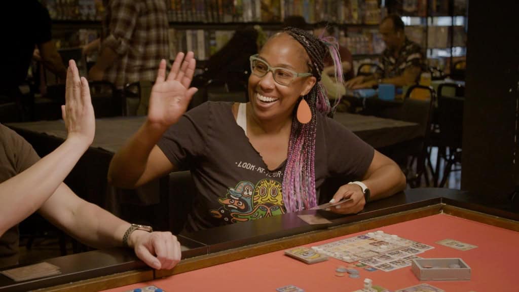 A woman with braided hair and glasses plays a card game, smiling and high-fiving someone across the table in a lively game store.