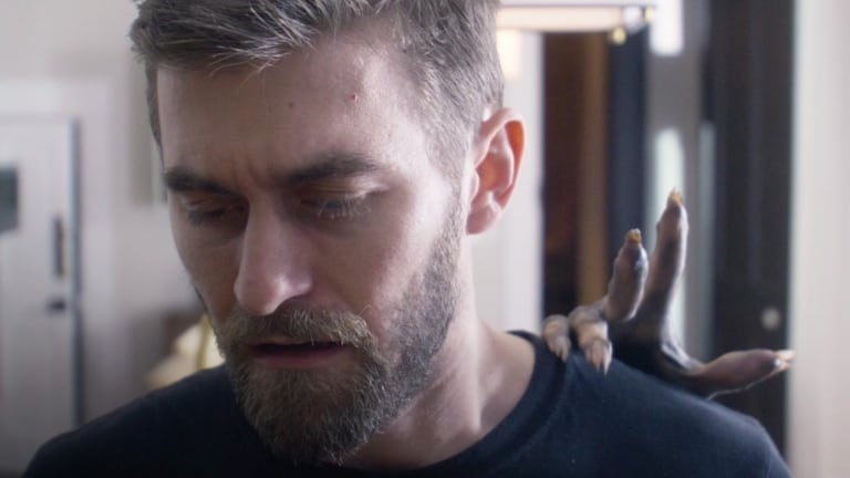 A man with a beard looks down thoughtfully as a small, brown lizard rests on his shoulder.