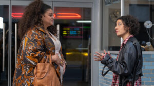 Two people are having a conversation indoors. One person is wearing a patterned coat and holding a brown bag, while the other person is wearing a black leather jacket over a red plaid shirt.