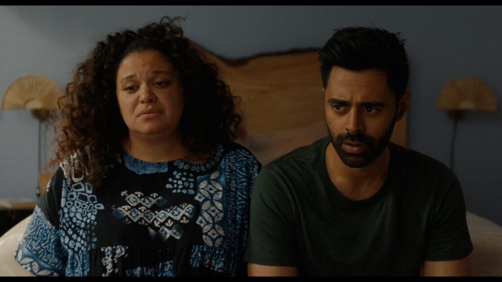A man and a woman sit on a bed, both looking serious and contemplative. The woman has curly hair and is wearing a patterned dress, while the man has short hair and is wearing a plain t-shirt.