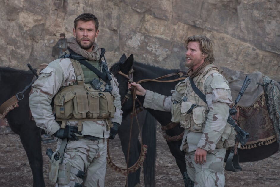 Two men in military gear standing with a horse in a rocky terrain, looking into the distance.