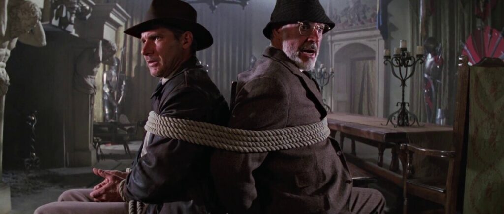 Two men wearing hats, tied back-to-back with rope, sit in a dimly lit room filled with antique furniture and statues.