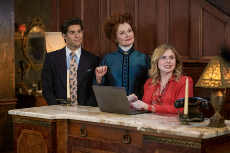 Three people standing by a marble counter in an ornate room, two women and one man, dressed in formal attire, appear engaged in a discussion.