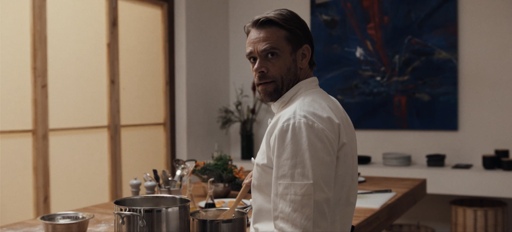 A man in a white chef coat stands in a kitchen, looking back over his shoulder. Cooking utensils and pots are on the counter in front of him.