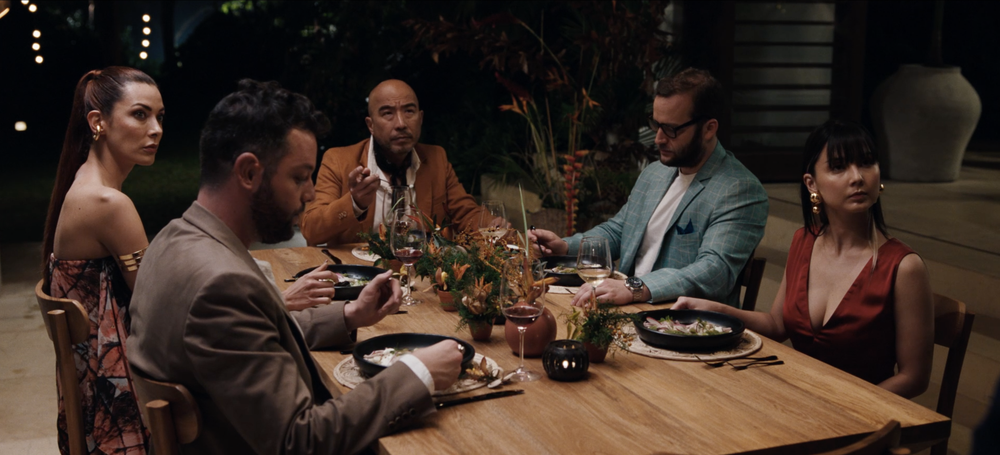 Five people are seated around a wooden table having a meal. The table is adorned with plants and decorative items, and the setting appears to be indoors or on a covered patio.