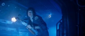 A person in a dimly lit space holding a flamethrower, looking alert with blue lighting and industrial surroundings.