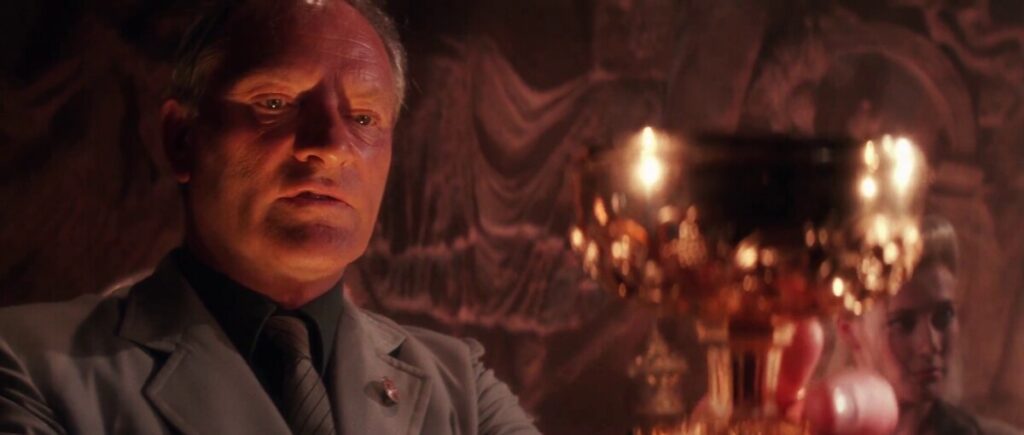 A man in a suit examines a golden goblet closely with a serious expression, while another person is blurred in the background. The surroundings are dimly lit with an elaborate carved wall.