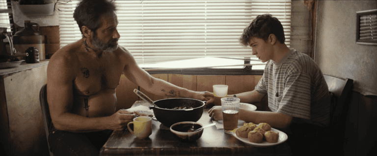A shirtless man and a young boy sit at a table inside a camper, eating and looking at each other, with a skillet and food items between them.