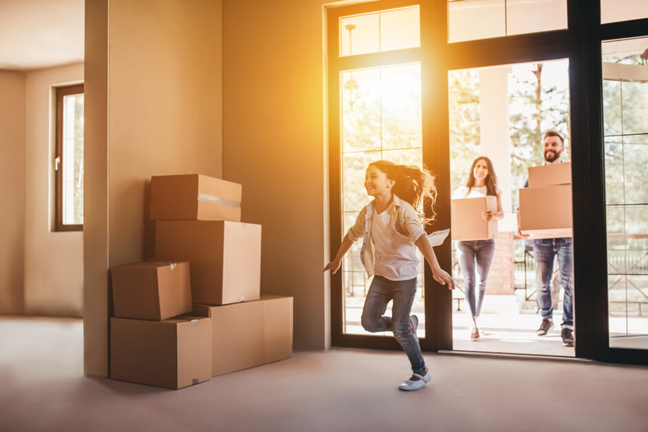 A young girl runs excitedly inside an empty room with stacked cardboard boxes, while two adults in the background carry more boxes through an open door.