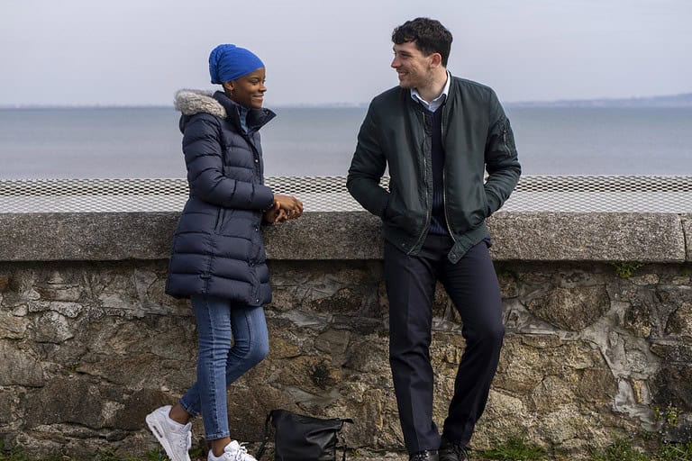 Two people, a woman in a blue beanie and a man in a green jacket, are smiling and talking while leaning on a stone wall by the sea.