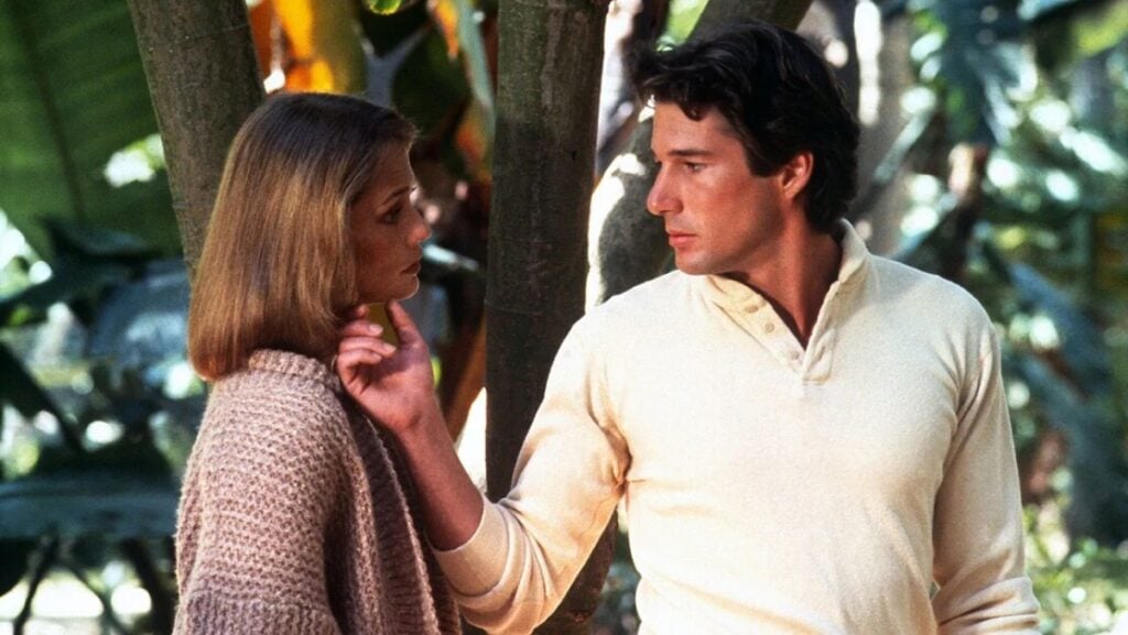 A man in a light-colored shirt touches a woman's face as she looks at him, standing outdoors near trees.