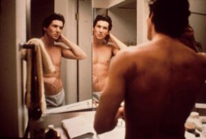 A shirtless man looks at himself in a bathroom mirror, one hand resting on his neck. Various toiletries are visible on the sink counter.