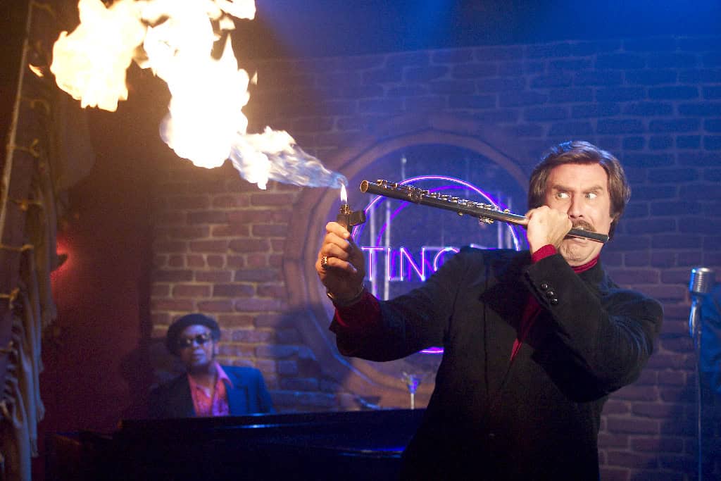A man performs a fire breathing trick by exhaling onto a flaming baton, with a musician playing piano in the background, in a dimly lit room resembling a club.