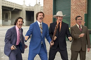 Four men dressed in 1970s outfits excitedly running together in an urban setting, two of them holding microphones.