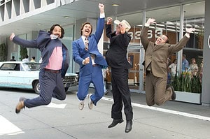Four men in business suits joyfully jumping in the air outside a building, with a vintage blue car and bystanders in the background.