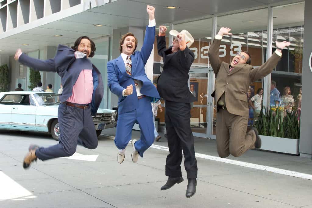 Four men in business suits joyfully jumping in the air outside a building, with a vintage blue car and bystanders in the background.