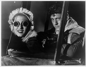A vintage black-and-white photo of a woman and man in a cockpit, wearing pilot goggles and helmets, with the woman smiling broadly.