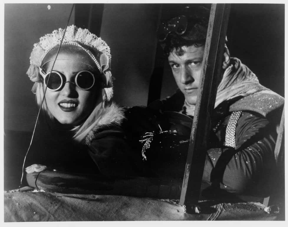 A vintage black-and-white photo of a woman and man in a cockpit, wearing pilot goggles and helmets, with the woman smiling broadly.