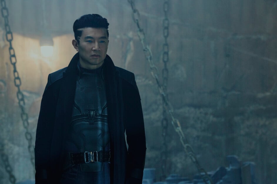 A man wearing a dark coat and armored suit stands in a dimly lit, industrial setting with chains hanging in the background.