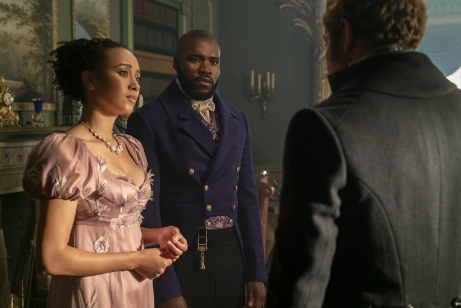 A woman and a man in 19th-century attire stand in a room facing another man with his back to the camera. The background includes vintage decor.