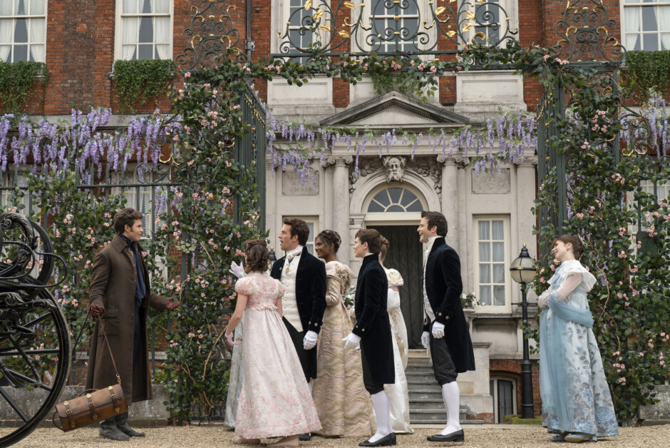 A group of people dressed in period clothing gather outside a grand building with flowers and ironwork.