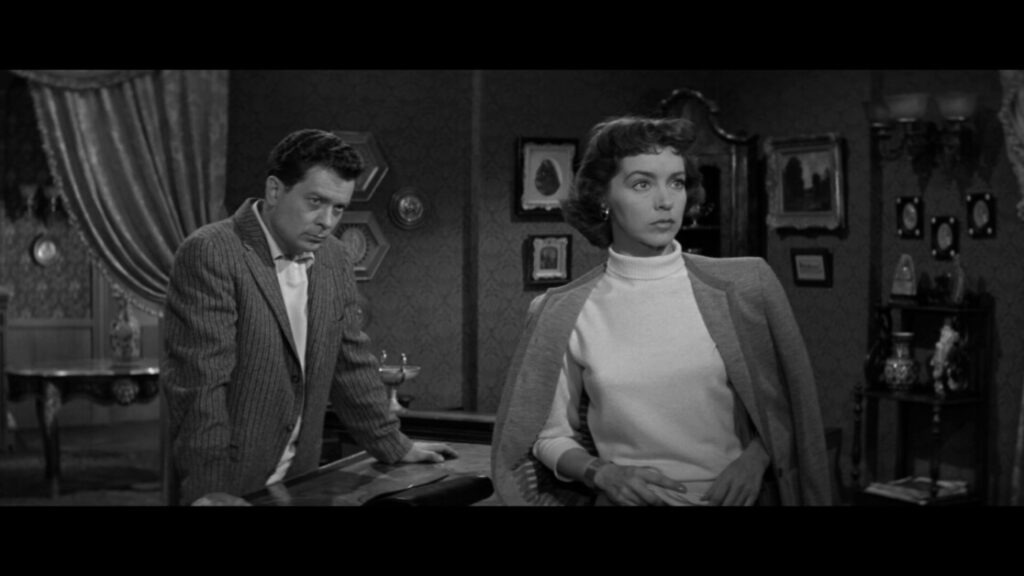 A man and a woman are in a vintage-styled room. The man is leaning on a desk while the woman stands with a pensive expression. The room features ornate decorations and framed pictures on the walls.