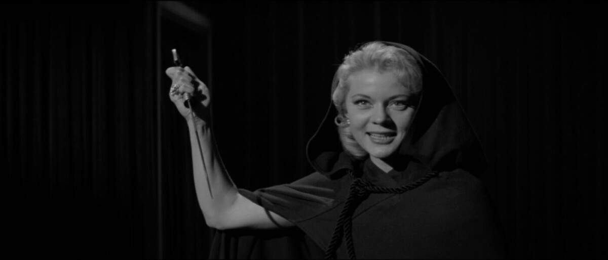 Woman in a hooded cloak smiles while holding up an object in her left hand against a dark curtain backdrop. The image is in black and white.