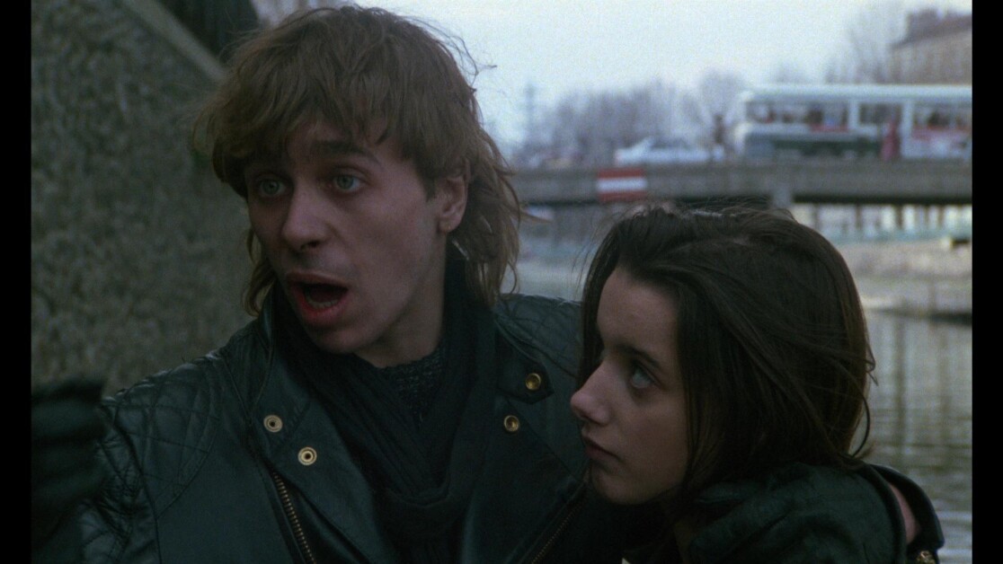 Two people standing near a waterway, both wearing dark clothing. The person with longer hair is speaking, while the other person looks off to the side, slightly leaning against them.