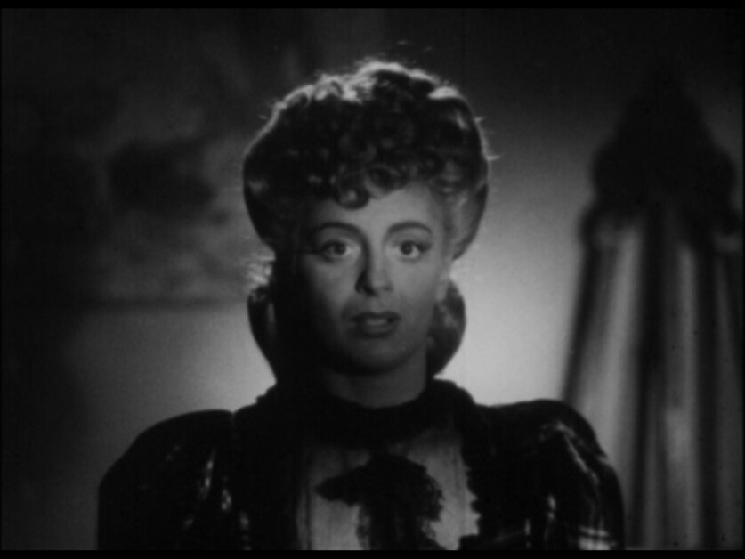 Black and white image of a woman with curly hair, looking directly at the camera, slight shadows cast over her face, wearing a dark top with a lace collar.