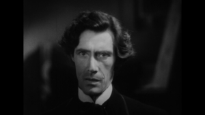 Black and white image of a man with striking facial features, wearing a dark suit and tie, looking intently at the camera with a serious expression.