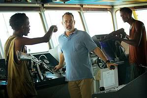 Two armed pirates confront a man inside a ship's navigation bridge, with tense expressions and a highly charged atmosphere.