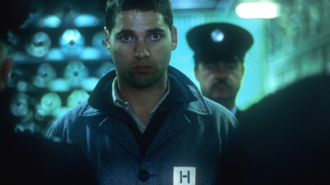 A man with a serious expression stands in a dimly lit room wearing a coat marked with an 'H', flanked by a uniformed officer in the background.