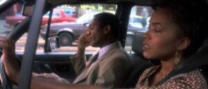 A man and woman are sitting in a car. The man is in the driver's seat, wearing a beige suit, and talking on the phone. The woman, in the passenger seat, looks annoyed or concerned.