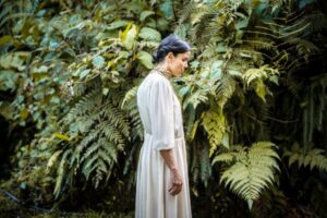 A woman in a white dress stands with her head slightly bowed in front of lush green foliage, with various types of ferns in the background.
