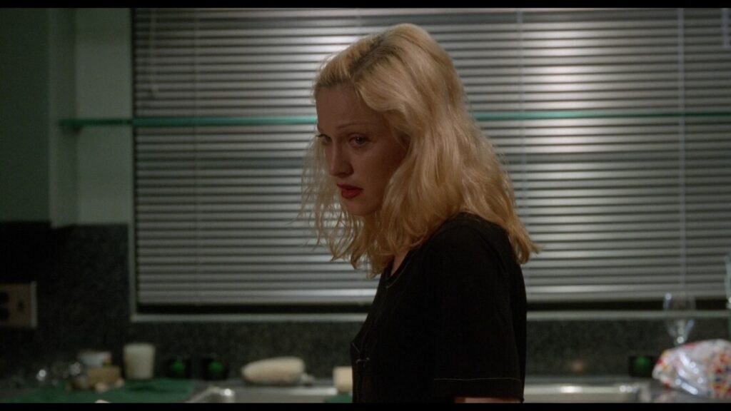 A woman with blonde hair and a black shirt stands in a kitchen with a serious expression, with blinds and various kitchen items visible in the background.