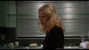 A woman with blonde hair and a black shirt stands in a kitchen with a serious expression, with blinds and various kitchen items visible in the background.